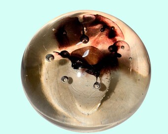 May include: A clear glass paperweight with a swirling, abstract design in shades of brown and black. The paperweight has a spherical shape and contains small air bubbles. The design resembles an ink blot.