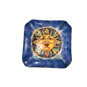 May include: Square ceramic dish with a blue textured background and a central sun face design. The sun face is yellow with a red mouth, surrounded by a gold and green border. The dish is likely for decorative use.