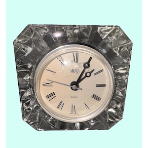 Puede incluir: Un reloj de cuarzo de cristal transparente y facetado con esfera color crema. La esfera del reloj presenta números romanos negros, manecillas negras y la marca "MIKASA". El reloj tiene forma cuadrada con bordes biselados.