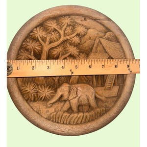 May include: A round, wooden, hand-carved decorative plate. The plate features a detailed relief carving of an elephant walking through a jungle scene, with trees, a hut, and mountains. The plate measures approximately 9 inches in diameter.
