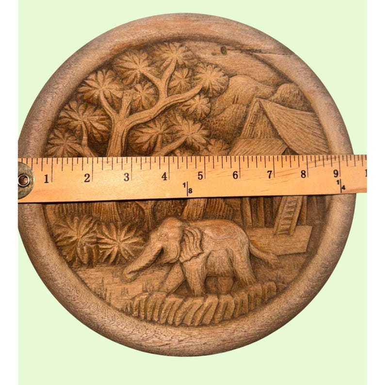 May include: A round, wooden, hand-carved decorative plate. The plate features a detailed relief carving of an elephant walking through a jungle scene, with trees, a hut, and mountains. The plate measures approximately 9 inches in diameter.