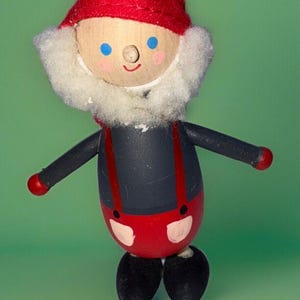May include: A wooden toy gnome with a red hat, white beard, and black and red clothing. The gnome has a painted face with blue eyes and a smile.