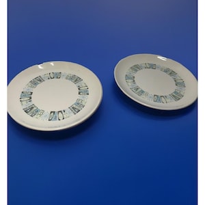 Op de afbeelding: Twee witte keramische borden met een decoratieve rand van blauwe, turquoise en zwarte geometrische vormen. De borden zijn rond en liggen op een effen blauw oppervlak. De borden zijn waarschijnlijk bedoeld om voedsel op te serveren.