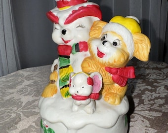 May include: A ceramic Christmas musical figurine of a snowman with a dog and a mouse. The snowman is wearing a red hat, a green scarf, and a yellow and black scarf. The dog is wearing a yellow hat and a red scarf. The mouse is wearing a red scarf. The figurine is on a white base with green holly leaves.