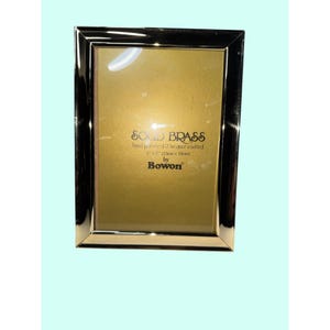 May include: A rectangular, polished brass picture frame with a gold-colored interior. The frame is labeled "SOLID BRASS" and "5 x 7" (13cm x 18cm) by Bowon."