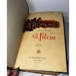Puede incluir: Libro abierto con el título "LA BOHEME" en una fuente decorativa. La portada presenta el texto "Opera in Four Acts BY G. PUCCINI". El libro es una partitura vocal en inglés e italiano, publicado por G. Ricordi & Co., Nueva York.