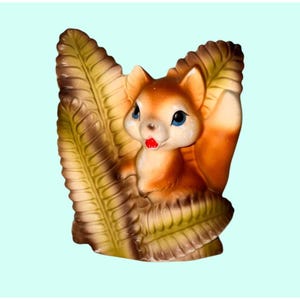 Op de afbeelding: Vintage keramieken eekhoornbeeldje met een roodbruin lichaam, witte borst en een pluizige staart. De eekhoorn heeft blauwe ogen en een rode mond en zit tussen twee grote, gestileerde varenbladeren in bruin- en groentinten.