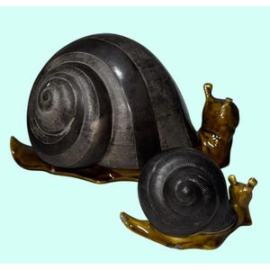 Può includere: Due sculture di lumache in ceramica. La lumaca più grande ha un guscio grigio scuro con strisce verticali e un motivo a spirale. La lumaca più piccola ha un guscio simile. Entrambe le lumache hanno corpi marroni e poggiano su una base marrone.