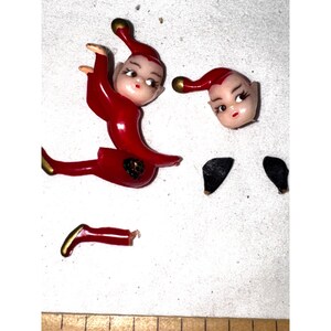 以下が含まれることがあります： 分解されたヴィンテージのクリスマスエルフの置物。エルフは主に赤色で、帽子と靴に金色のアクセントがあります。エルフの顔は薄い色で、顔の特徴が描かれています。エルフの黒い靴と頭は体から分離されています。
