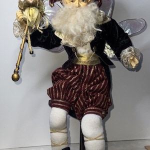 May include: A whimsical fairy doll with a black velvet jester hat, a white beard, and red and gold striped pants. The doll has iridescent wings and is holding a golden wand with bells.