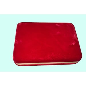 May include: A rectangular, red velvet jewelry box with rounded corners. The box has a gold-colored trim around the edges and a soft, plush texture. The background is a light blue.