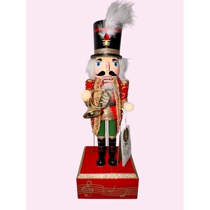 May include: A decorative wooden nutcracker soldier in a red and gold uniform, holding a brass instrument. The nutcracker has a black top hat with a feather, white hair and mustache, and is standing on a red base with musical notes.