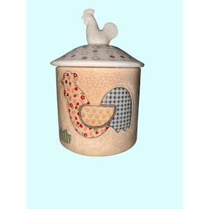 May include: A ceramic jar with a lid, featuring a rooster design. The jar has a beige, crackled finish with a rooster illustration in floral and gingham patterns. A white rooster figurine tops the lid, which has a floral pattern.