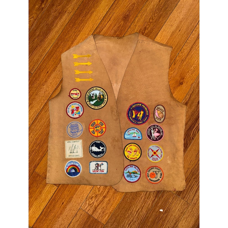 May include: Tan vest with multiple colorful patches. Patches include images of a rainbow, a whale, arrows, and text. The vest is made of a suede-like material and has a V-neck design.