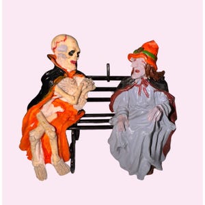 Puede incluir: Figuras de Halloween de un esqueleto y una bruja sentados en un banco negro. El esqueleto lleva una capa naranja y negra, mientras que la bruja viste un vestido gris y un sombrero naranja. Las figuras son detalladas y coloridas.