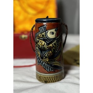 May include: A small, cylindrical ceramic container with a detailed fish design. The container features a brown background with a blue and gold fish illustration. It has a dark blue rim and a woven base, with a brown cord handle.