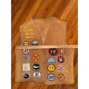 May include: Tan vest with multiple colorful patches. The vest has a V-neck and is made of a suede-like material. Patches include arrows, a whale, a rainbow, and various activity emblems. The vest measures approximately 24 inches across.