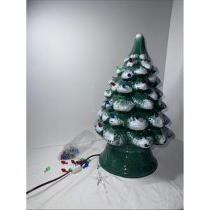 May include: A ceramic Christmas tree in a glossy green finish, decorated with white snow-like details and colourful lights. The tree has a star-shaped topper and sits on a matching green base. A bag of extra lights and a power cord are included.