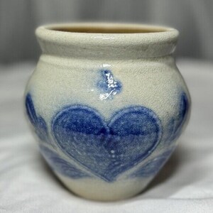 May include: A small white ceramic pot with a blue heart design. The pot has a rough, textured surface and a slightly rounded shape.