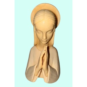 May include: A carved wooden bust of a woman with a halo, hands clasped in prayer. The figure has a serene expression, with closed eyes and a gentle downward gaze. The sculpture is in a light beige colour.