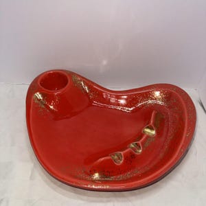 Puede incluir: Un cenicero de cerámica roja con forma de riñón. El cenicero tiene un soporte circular y tres reposacigarrillos. Hay motas doradas dispersas por la superficie. Un artículo vintage.