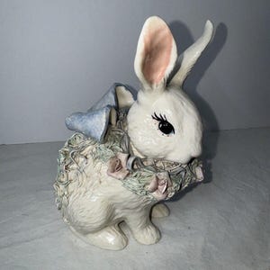 May include: A white ceramic bunny figurine with a blue and green wreath around its neck. The bunny has pink ears and black eyes.