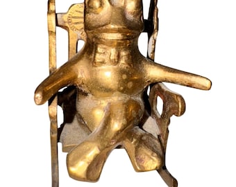 May include: A brass frog figurine seated in a rocking chair. The frog has outstretched arms and is wearing a bow tie. The chair has a sunburst design on the back.