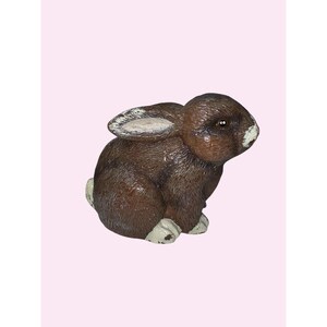 May include: A brown rabbit figurine with white accents on the ears, nose, and feet. The bunny is in a seated position, with its ears perked up. The figurine has a textured surface and is set against a light pink background.
