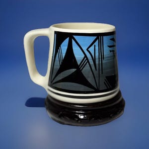 May include: A white ceramic mug with a black and blue geometric design. The mug has a handle and sits on a wooden base.