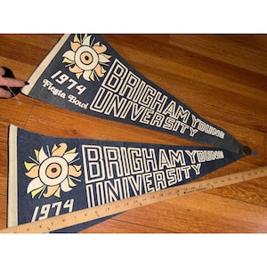 以下が含まれることがあります： 黄色と白のスタイライズされた太陽のグラフィック、「1974 Fiesta Bowl」、「BRIGHAM YOUNG UNIVERSITY」という白い文字が特徴の濃い青色のフェルト製ペナント2枚。木製の定規により、下のペナントの長さが約71cmであることがわかります。