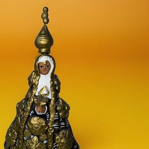 Op de afbeelding: Een klein, goud geverfd beeldje van een vrouw die een witte sluier en een gouden gezoomde jurk draagt. Het beeldje staat op een gele ondergrond.