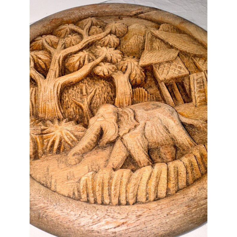 May include: A carved wooden relief depicting an elephant walking through a forest scene. The relief is circular, with detailed carvings of trees, foliage, and a small structure. The wood has a natural, light brown color.