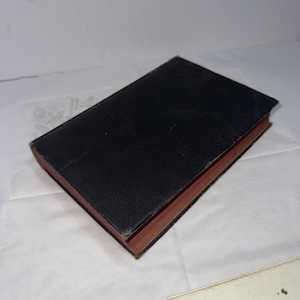 May include: A closed, rectangular book with a black cover and red-toned edges. The cover has a textured appearance. The book is resting on a white surface.