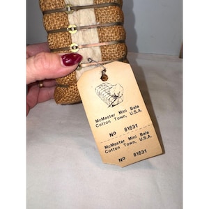 May include: A miniature cotton bale wrapped in brown burlap and secured with thin metal bands. A beige paper tag is attached, featuring a line drawing of a bale and the text: McMaster Mini Bale, Cotton Town, U.S.A., No 81631.