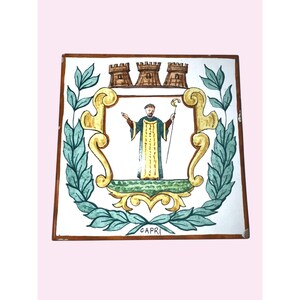 May include: A square ceramic tile featuring a hand-painted illustration of a figure in robes holding a staff, centered within a yellow scrollwork frame. Above the frame are three brown towers, and the word CAPRI is written at the bottom.