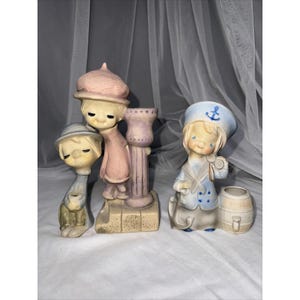 Może przedstawiać: Dwie ceramiczne figurki przedstawiające stylizowane postacie. Jeden element ukazuje dwie postacie w różowo-beżowych strojach przy kolumnie. Drugi element przedstawia postać w niebieskim stroju marynarskim z czapką z kotwicą, stojącą przy beczce.