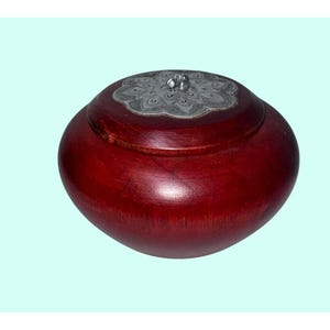 May include: A round, wooden box with a deep red finish. The lid is adorned with a silver-colored, floral-patterned metal plate. The box has a smooth, polished surface, and the lid has a small, decorative handle.