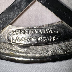 May include: Close-up of a silver-toned metal object with the text "ANNIE KAIT... BAREFOOT DREAMS" embossed on it. The object has a textured surface and a triangular shape at the top.
