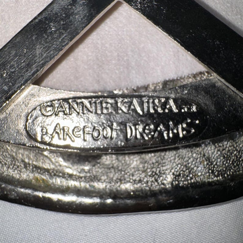 May include: Close-up of a silver-toned metal object with the text "ANNIE KAIT... BAREFOOT DREAMS" embossed on it. The object has a textured surface and a triangular shape at the top.