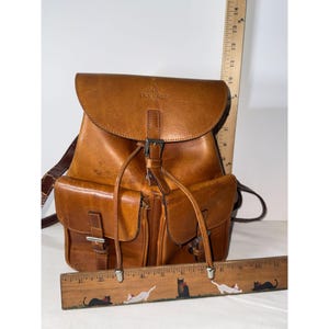 May include: A tan leather backpack with a top flap, two front pockets, and silver-toned metal buckles. A vertical ruler shows the bag is approximately 11 inches tall, and a horizontal ruler shows it is approximately 11 inches wide at the base.