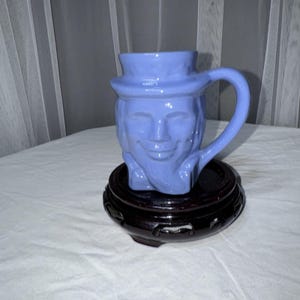 Könnte beinhalten: Hellblaue Keramiktasse mit einem lächelnden Gesicht, Henkel und hutartigem Rand. Die Tasse steht auf einem dunkelbraunen, verzierten Holzständer. Die Höhe der Tasse beträgt ca. 13 cm.