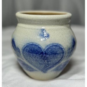 May include: A small, off-white ceramic jar with a wide opening and a slightly textured surface. The jar is decorated with large, blue heart-shaped designs. The interior of the jar is a light brown color.