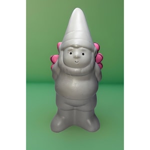 Puede incluir: Figura de gnomo de jardín plateado con un gorro puntiagudo blanco y detalles de flores rosas. El gnomo tiene una cara sonriente y está de pie. El fondo es de color verde liso.