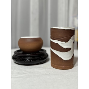 Puede incluir: Dos piezas de cerámica: un cuenco pequeño y redondo de color marrón con borde blanco, sobre un soporte de madera negro y ornamentado, y un jarrón alto y cilíndrico marrón con un diseño blanco en forma de ola. Para decoración del hogar.