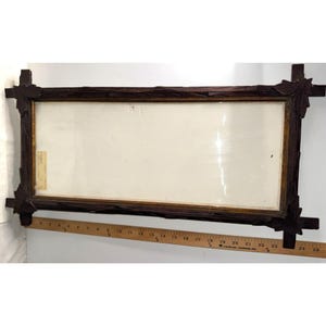 May include: A rectangular dark brown wooden picture frame with carved leaf details at each corner. A wooden ruler is positioned along the bottom edge, showing the frame length is approximately 24 inches. The frame contains a blank white insert.
