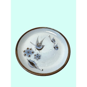 May include: A round ceramic plate with a light blue glaze and a dark brown rim. The surface features hand-painted illustrations of a small bird in flight, two blue butterflies, two stylized floral blossoms, and a single leaf in shades of blue and brown.