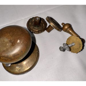 May include: Antique brass door knobs and hardware. The set includes two large, round knobs, and several smaller pieces with decorative detailing. The hardware has a warm, aged patina, suggesting vintage or salvaged origins. The pieces are arranged on a white surface.