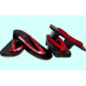 May include: Black geta sandals with red straps. The wooden sandals have a glossy finish and are decorated with floral patterns. The traditional Japanese footwear is displayed on a light blue background.