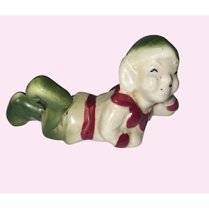May include: A ceramic figurine of a reclining elf with green pants and a green cap. The elf's cream-colored body is adorned with red stripes and accents. The figurine is lying down, with its head resting on its hand.