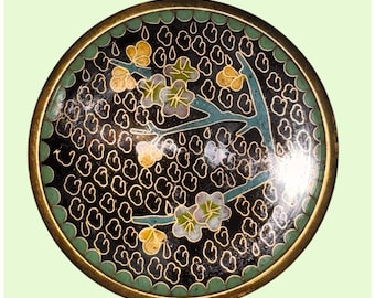 May include: Circular cloisonné ornament with a black base and a floral motif. It has a gold-coloured rim, a green border, and a branch with white and yellow flowers. The background features a repeating pattern of gold-coloured shapes.